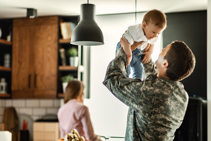 Happy baby boy having fun with his military dad at home. Mother is in the background.