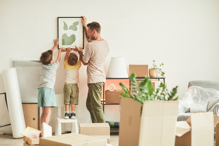 Full length portrait of happy father with two sons hanging pictures on wall while moving in to new home, copy space